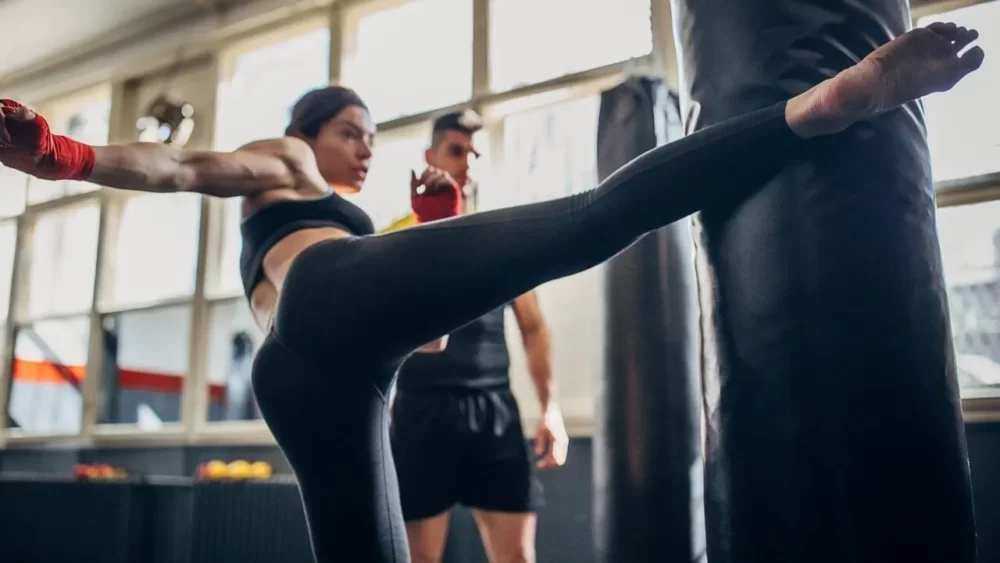 Image-of-a-woman-practicing-a-right-middle-kick-with-a-trainer-and-a-sandbag